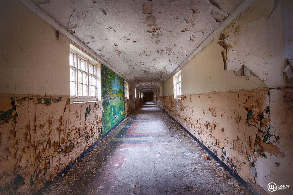 Long, abandoned hospital hallway with peeling paint, natural light, and a faded mural of a landscape scene. Dirty floor, damaged walls, and a sense of decay.
