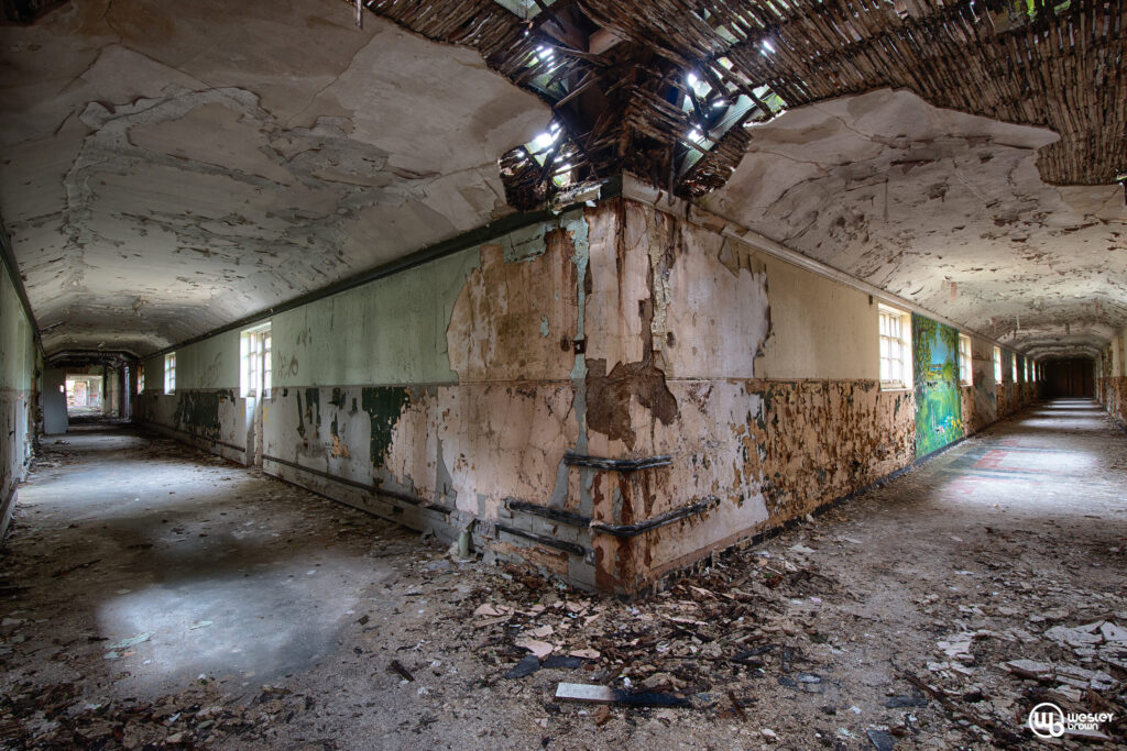 A long, decaying hallway splits into two paths, with peeling paint on walls, broken ceiling, and debris covering the floor.