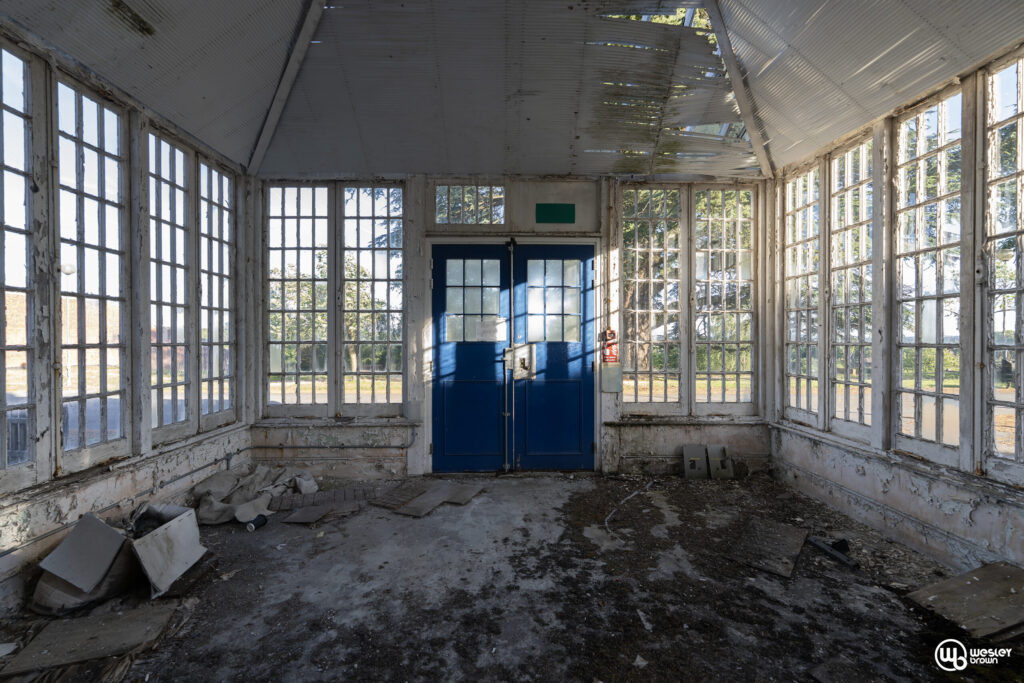 Broadmoore Hospital_30 copy Interior of an abandoned room with peeling paint and broken windows, bright daylight shining through.