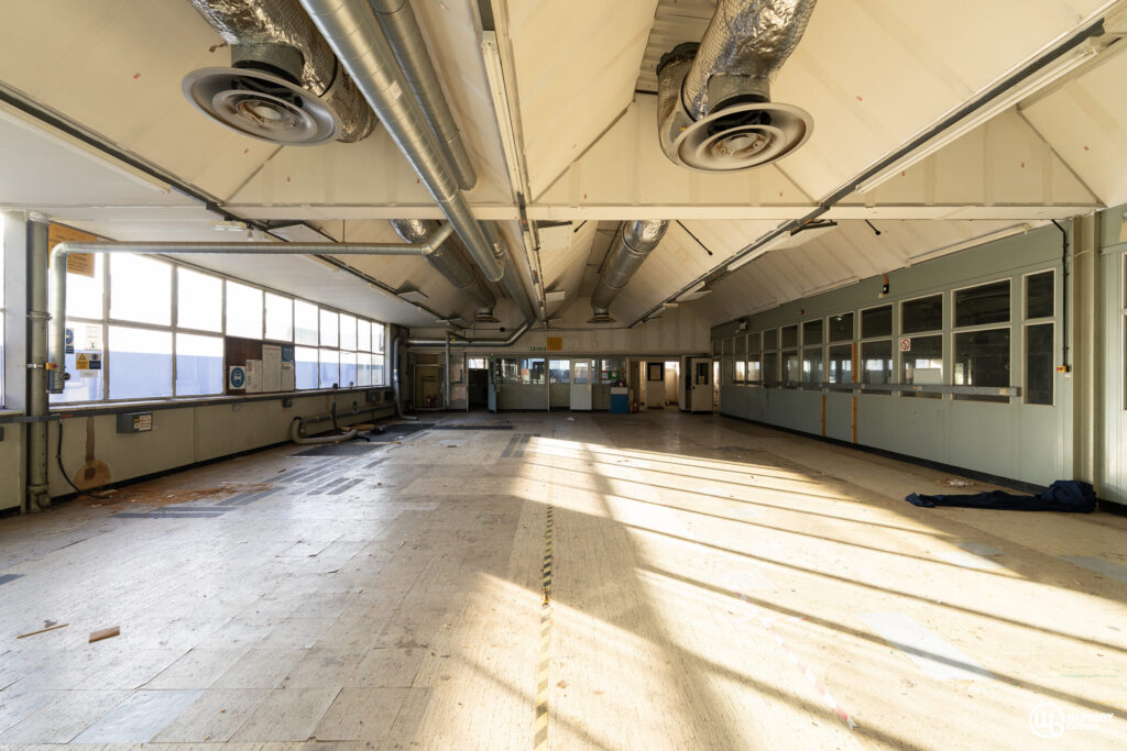 Broadmoore Hospital_31 copy Interior of an empty industrial building with exposed ducts and large windows.